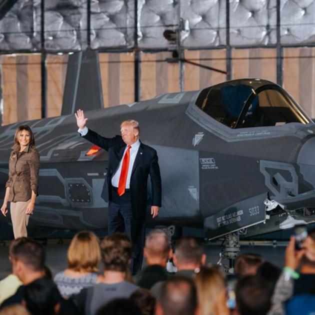 Trump, heavyset man in suit with red tie waving next to wife Melania, shapely woman with long brown hair standing in front of a military plane with people in foreground facing him
