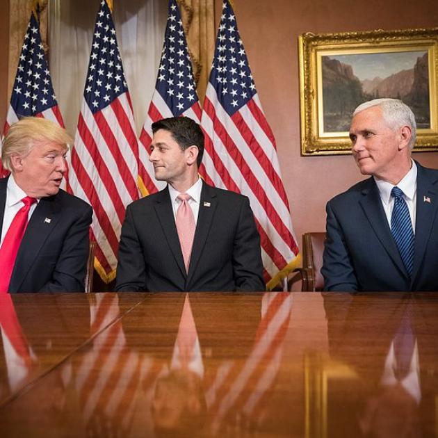 Trump, Ryan and Pence at a table looking at each other with flags in the back