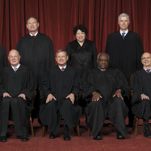 Photo of men and women posing and facing the camera all wearing long black robes in front of a dark red curtain