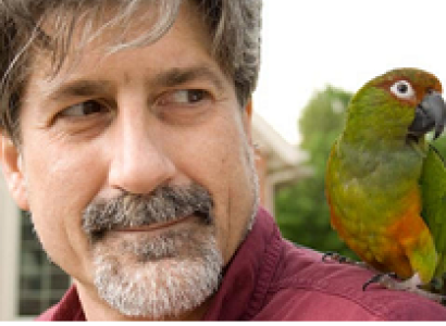White man with grayish hair and beard with green parakeet on his shoulder