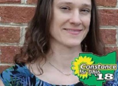 Young white woman with brown curly hair smiling standing in front of a brick wall with the words Constance for Ohio '18 in the right side bottom