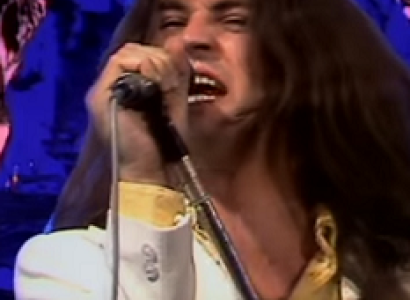 Long white man rock singer belting out a song into a mic with a blue and pink psychedelic background