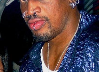 Black man in a baseball cap with lots of facial rings in lips, nose and ears wearing a blue shirt