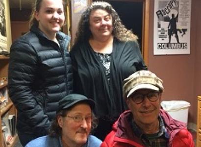 Two women, one young with hair pulled back and the other older with long brown curly hair standing behind two men kneeling, both wearing hats with brims and wire rimmed glasses, everyone smiling