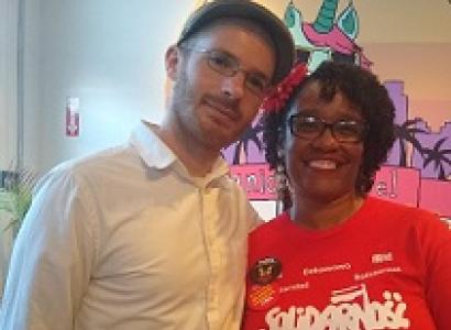 Young white man with facial hair, glasses and a hat posing with his arm around a black woman in a solidarity T-shirt
