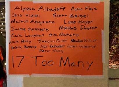 A sign on orange paper taped to a brown wall listing all the names of people killed in the recent Parkland Fla school shooting and the words 17 too many at the bottom 