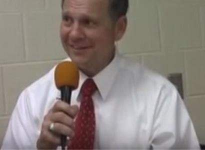 White man with brown hair, white shirt and red tie talking into a microphone