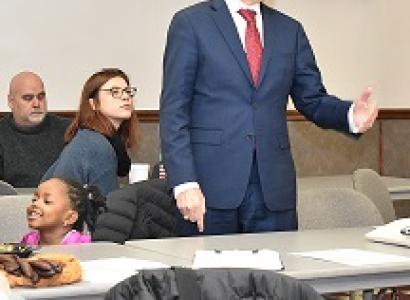 Middle aged white man with some dark and some gray hair speaking while gesturing to the right wearing a suit, people listening to him
