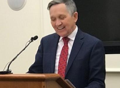 Older white man in suit with red tie at a brown podium looking down, smiling and talking into a mic