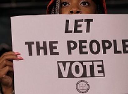 Black person's face with a red hat on their head peering over a sign that they are holding that says Let The People Vote