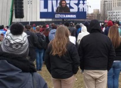 Backs of people standing watching a speaker on a screen