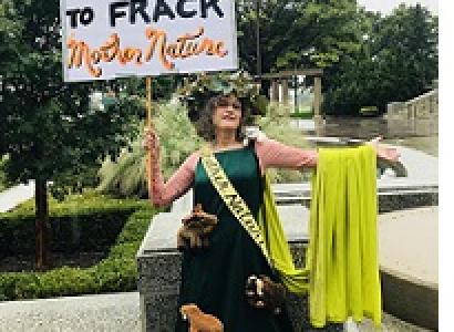 Woman in a long green dress with a sash and a cape and crown of flowers with a sign saying It's not nice to frack mother nature