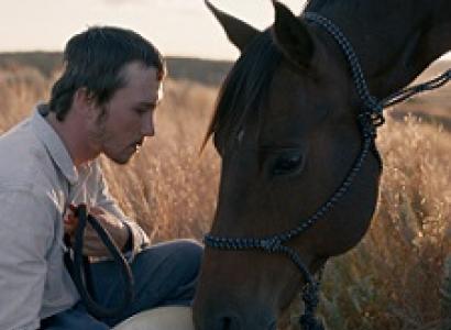 Young man with brown hair looking sideways and a big brown horse leaning its head toward his lap