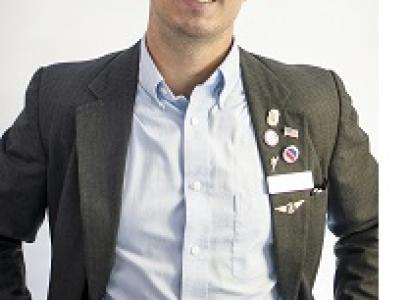 Young white man with dark rimmed glasses and a suit with lots of political buttons and a big smile