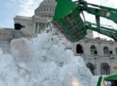 Big white stately building with round top and lots of windows behind a huge green truck with a big arm and huge shovel putting a ton of white snow in a big pile in front