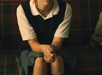 Young white woman in a white blouse, vest and hair pulled back sitting on a couch with hands clasped looking irritated
