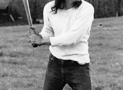Black and white photo of a young man with long flowing brown hair with a headband across his forehead holding a baseball bat as if he's about to swing