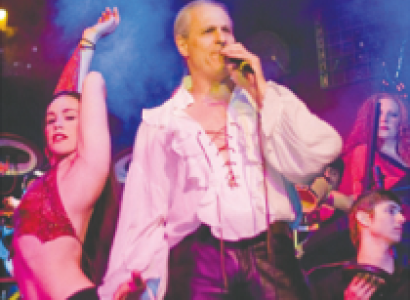 Middle aged white man with balding head wearing a blousy tie-front white shirt with tight shiny dark pants with a woman dancing sideways wearing a red bikini top and low-rider black pants and others dancing behind