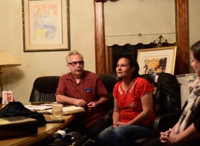 Gray haired man with black-rimmed glasses sitting at a table looking at a dark haired woman who is speaking also sitting in a chair