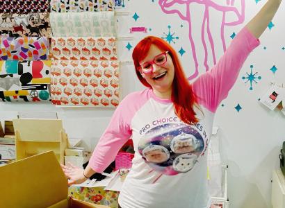 Woman with red hair and pink glasses holding up her hand like waving hello in a room with a lot of arts and crafts supplies