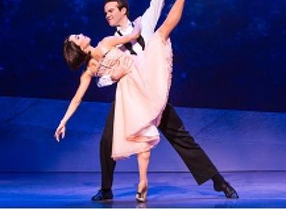 White man with dark hair in white shirt and black pants dancing with a white woman with short dark hair in a pick dress, he is leaning over as he holds her as she is kicking her leg high up in the air 
