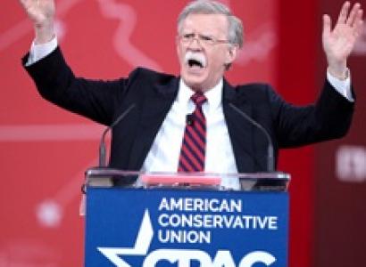Older man with gray hair and long white mustache standing at a podium with his mouth  open waving both hands in the air and the words American Conservative Union on the podium