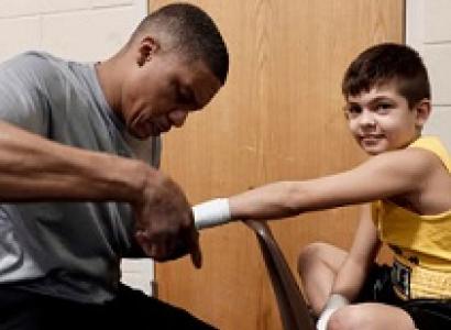 Black man in gray shirt taping up the hand of a young white boy in a yellow tank top