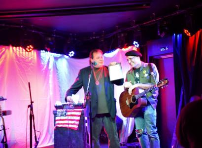 Two men on a stage, one with a guitar, one with a plaque in his hand