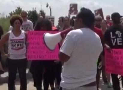 Man with brown hair and white shirt with back to camera holding a bullhorn surrounded by protestors