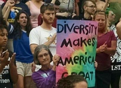 Person holding sign in crowd of people with fists in air, reading Diversity Makes America Great against tie-dyed background
