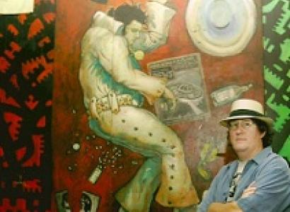 White man in blue shirt with straw hat with arms folded standing in front of huge mural of Elvis Presley