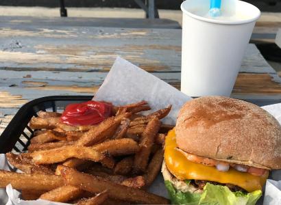 A tray on a wooden table outside with a white cup and blue straw, a bunch of french fries and a burger with visible lettuce and cheese