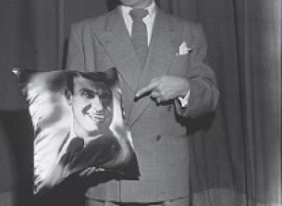 Black and white photo of white man in a suit pointing to a picture of himself in his hand