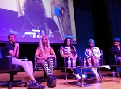 Stage with large screen with a black trans woman speaking and five black women in chairs below