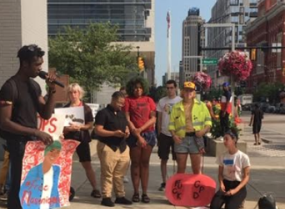 Young black man speaking into a mic to a crowd of people outside with signs