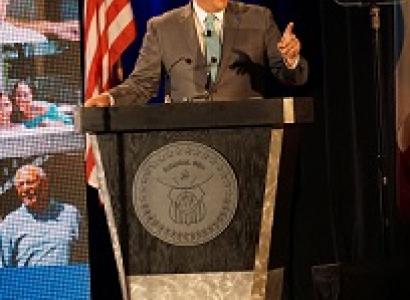White middle-aged man in a brown suit at a podium pointed and speaking with an U.S. flag and a screen with pictures behind him