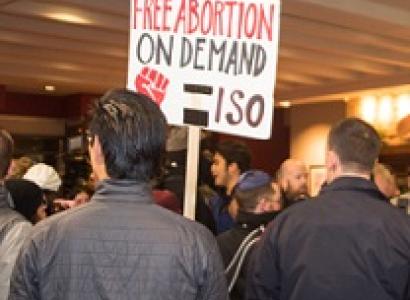 Backs of two men, one with a cop looking uniform on and the other with a sign that says Free Abortion on Deand ISO with a red fist