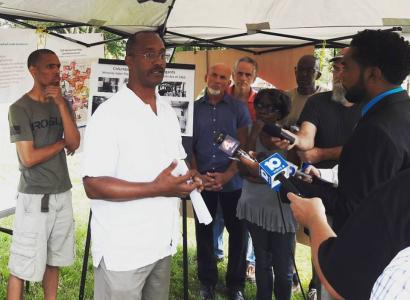 A press conference with a black man in a suit holding a mic for another black man with glasses and a white shirt to talk into and other people standing around watching
