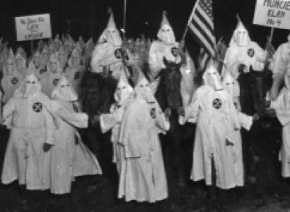 People in white robes with pointy top hats in a circle outside at night with a flag