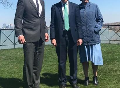 Three people standing outside on the grass with  body of water behind them, a tall white man in a suit with gray hair, and shorter white man in a suit and a black woman with glasses