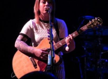 Young white woman in a white t-shirt playing a guitar and singing at a mic with a black background
