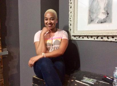 Young Black woman smiling and posing with her hand to her chin next to a painting