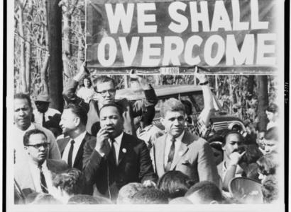 Black and white photo of Martin Luther King Jr. and others marching under sign that says We Shall Overcome