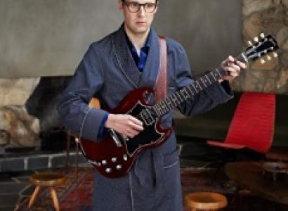 Young geeky guy with big black rimmed glasses wearing a bathrobe holding an electric guitar in a room with two wooden stools
