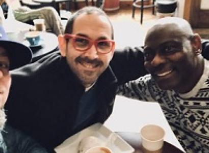 A balding white guy with red framed glasses and facial hair smiling sitting at a table with his arm around a bald black man in a white and black designed sweater also smiling