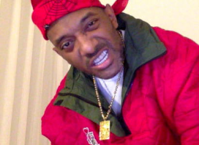 Black man in red jacket with red doo rag scarf on head with gold necklace, making a sneering face