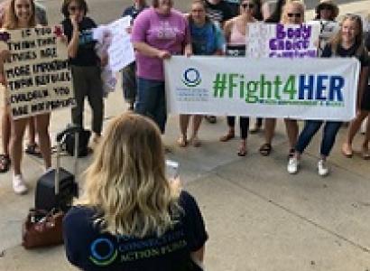 A back of a woman in the foreground speaking outside to a group of women facing her holding signs saying #Fight4Her