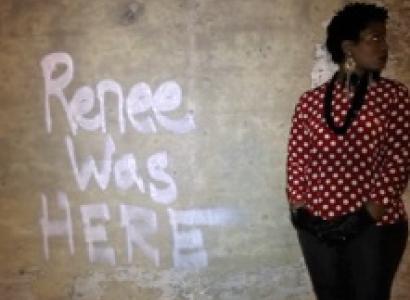 The words Renee was HERE spray painted on a brown wall and a black woman with short hair and a red and white shirt with a black necklace and black pants stands against the wall looking to the right