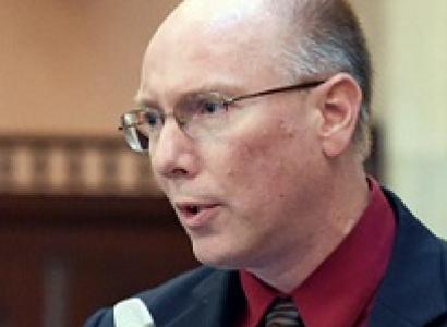 Balding white guy with sire rimmed glasses, a blue suit coat and maroon shirt and striped tie holding a silver microphone talking into it