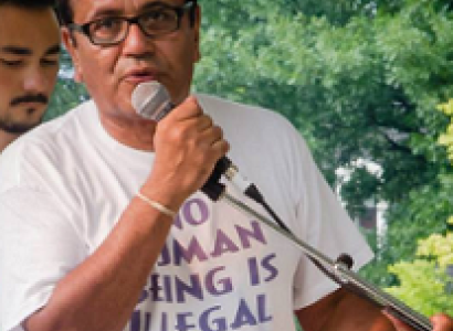 Latino man in T-shirt that says No Human Being is Illegal, talking into a mic